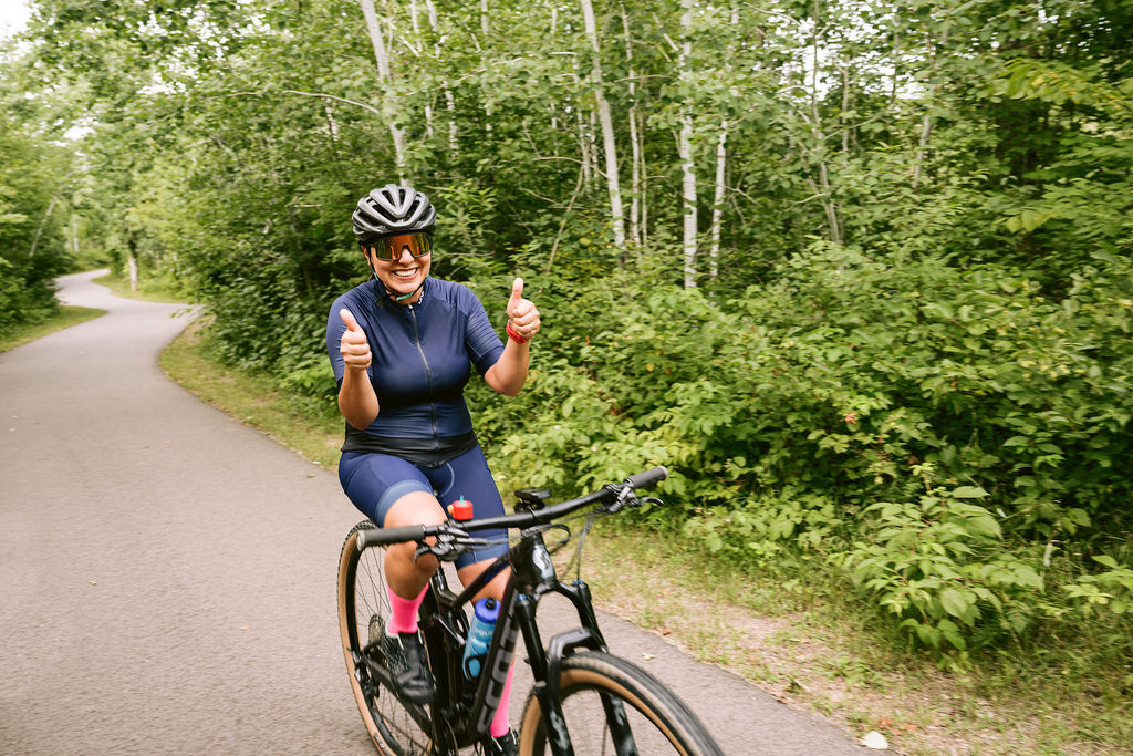 Woman on bike on paved trail. She's wearing a blue biking outfit and smiling and giving two thumbs up to the camera.