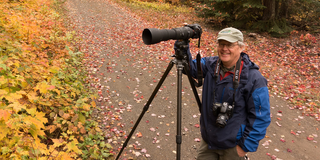 Man with an SLR camera standing on a trail on a fall day.