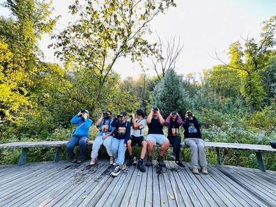 Group of people sitting on a bench outside with binoculars to their eyes looking up.