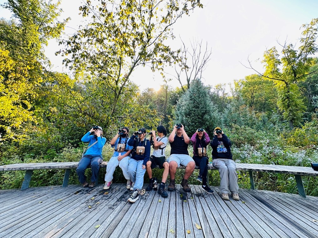Group of people sitting on a bench outside with binoculars to their eyes looking up.