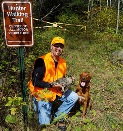 a grouse hunter with a dog and a bird he harvested by a Hunter Walking Trail sign and trail