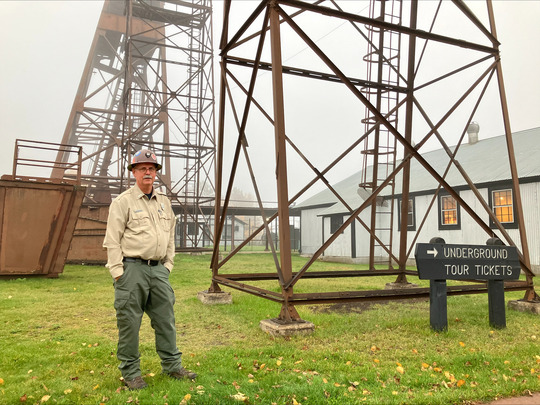 Park ranger in uniform, wearing a hard hat and posing by mine structures at a park.