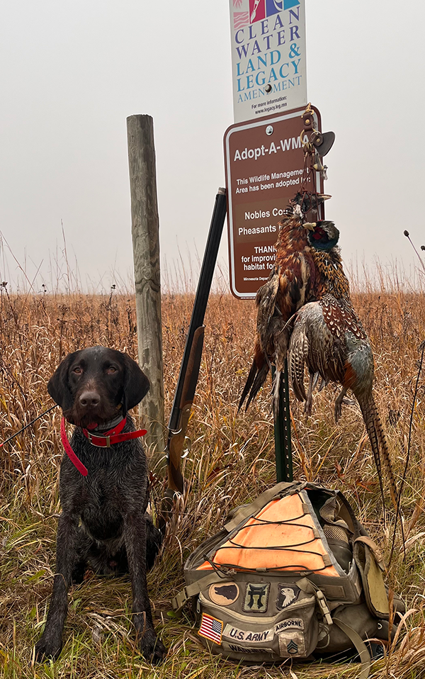 pheasants on a post with a dog by an adopt a WMA sign and Legacy amendment sign, and a U.S. Army Airborne backpack