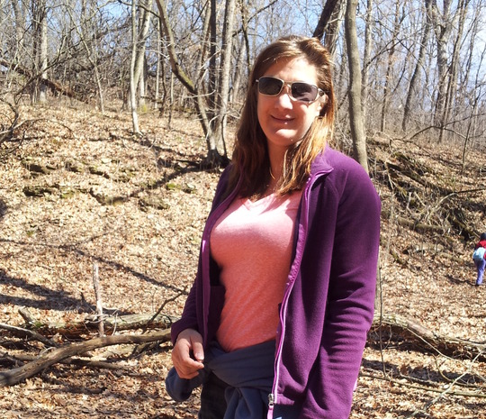 A woman standing in a fall forest, with people walking in the background.