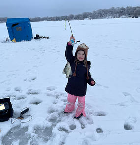 girl with a crappie she caught ice fishing, she's holding up the fish with it on the line while standing on the ice covered lake 
