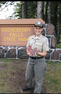 Person in park uniform holding red leaves and posing by Gooseberry Falls State Park sign.