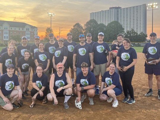 A group of people in matching t-shirts, standing on a softball field with a sunset in the background