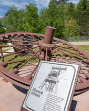 A huge iron wheel lays on its side with a descriptive plaque in front of it.