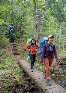 Two people walk along a small wood bridge in a forest with large camping backpacks on. 