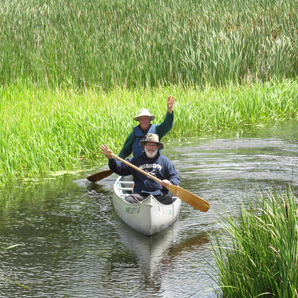 Two people wave and smile as they canoe down a river. 