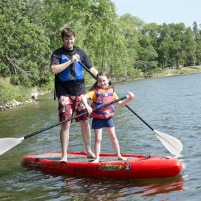 A parent and child paddleboard together on a lake during summer. 