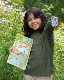 A young child holds out a patch with a fox on it and a coloring book that says Junior Ranger while smiling. 