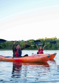 A child and adult smile while kayaking and holding their paddles above their heads. 