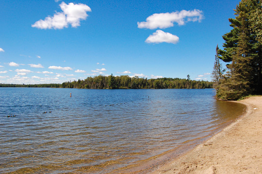 A calm lake stretches out into the horizon. In the foreground is a sandy beach. 