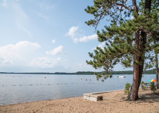 A beach with swimming area roped off stretches out into a calm, blue lake. 