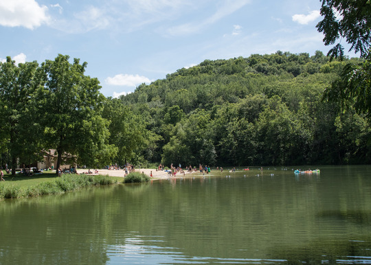 A small sandy beach at a lake is surrounded by lush green trees and a grassy area. 