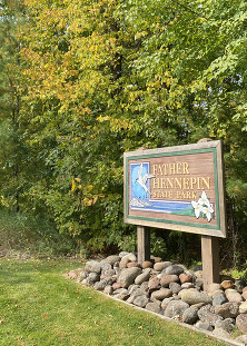 A wood painted sign next to a forested area reads Father Hennepin State Park. 