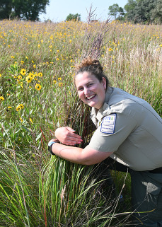 A person in a ranger uniform smiles broadly while hugging a clump of prairie grass. 