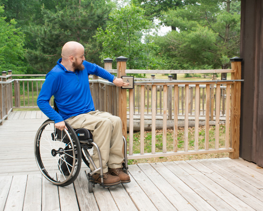 A person in a wheelchair pushes a button on an outdoor ramp to open the building's door. 