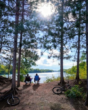 Two people sit on chairs looking at a lake surrounded by pine trees with bikes in the foreground. 