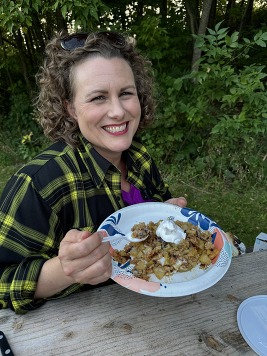 A woman smiles broadly while eating a plate of apple crisp at a state park picnic table. 