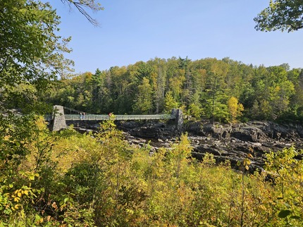 A historic stone swinging bridge stretches across a river, surrounded by yellowing trees. 