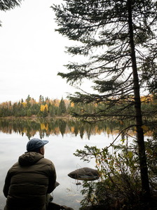 A person sits by a calm lake and looks out at the forest surrounding the water. 