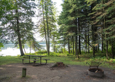 A campsite next to a calm lake with a campfire ring and picnic table.