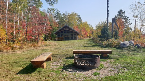 A campfire site in the foreground with a little cabin behind it, all surrounded by gorgeous fall forest. 