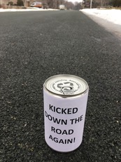 Can in road with label "Kicked down the road again!"