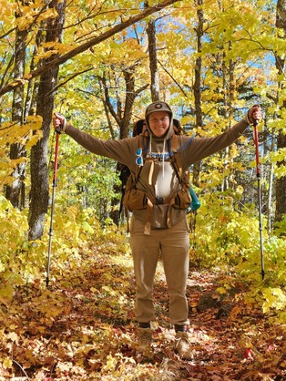 A person looks joyfully at the camera while hiking through an orange fall forest. 