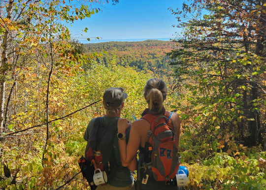 Two people look out at a forest of bright colors stretching to Lake Superior. 