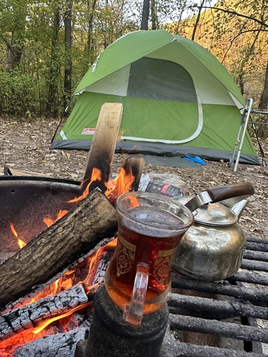 A cup of tea and bonfire in the foreground with a tent and fall forest in the background. 