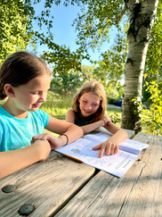 Two young children sit at a picnic table outside and look at a coloring book.