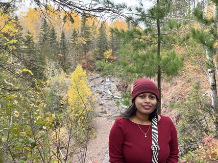 A person wearing red smiles with a fall forest behind them. 