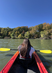 A person kayaks on a calm lake. The trees surrounding the lake have changed colors for fall.