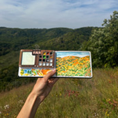 A hand holds up a mini water color painting of a fall landscape. 