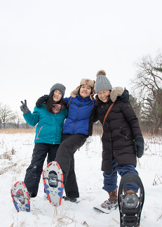 Two children and a parent smile and wink at the camera while holding up a snowshoe each. 