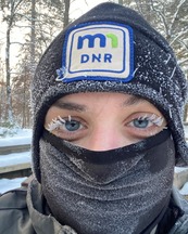 A person wearing a neck gaiter and hat has frozen eye lashes. 