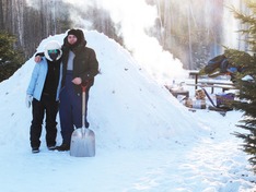 Two people stand in winter outdoor gear with a bonfire and snow structure behind them. 