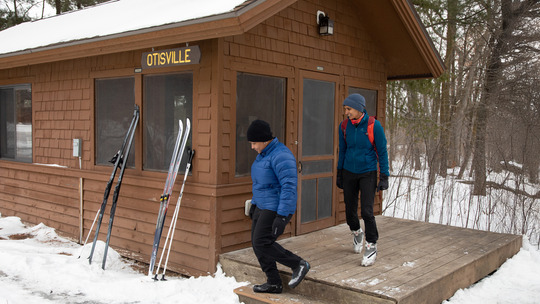 Two people exit a camper cabin in winter. Their skis are leaning up against the outside of the cabin. 