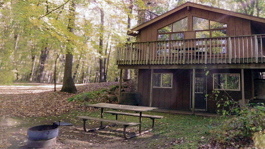 A large guesthouse sits back in a wooded area with picnic table and fire ring out front. 