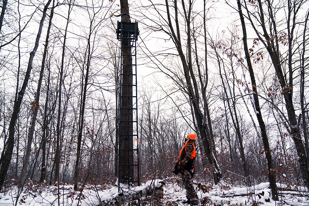 firearms deer hunter in blaze orange standing by a ladder stand in snowy woods