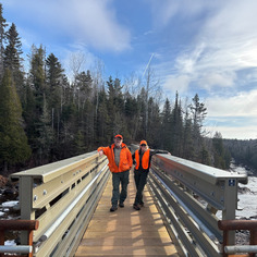 Two people stand on a new bridge and smile at the camera in orange vests. 