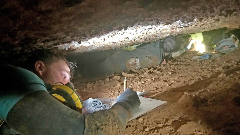 A researcher lies flat writing on a clip board in a very narrow crevice in a cave. 