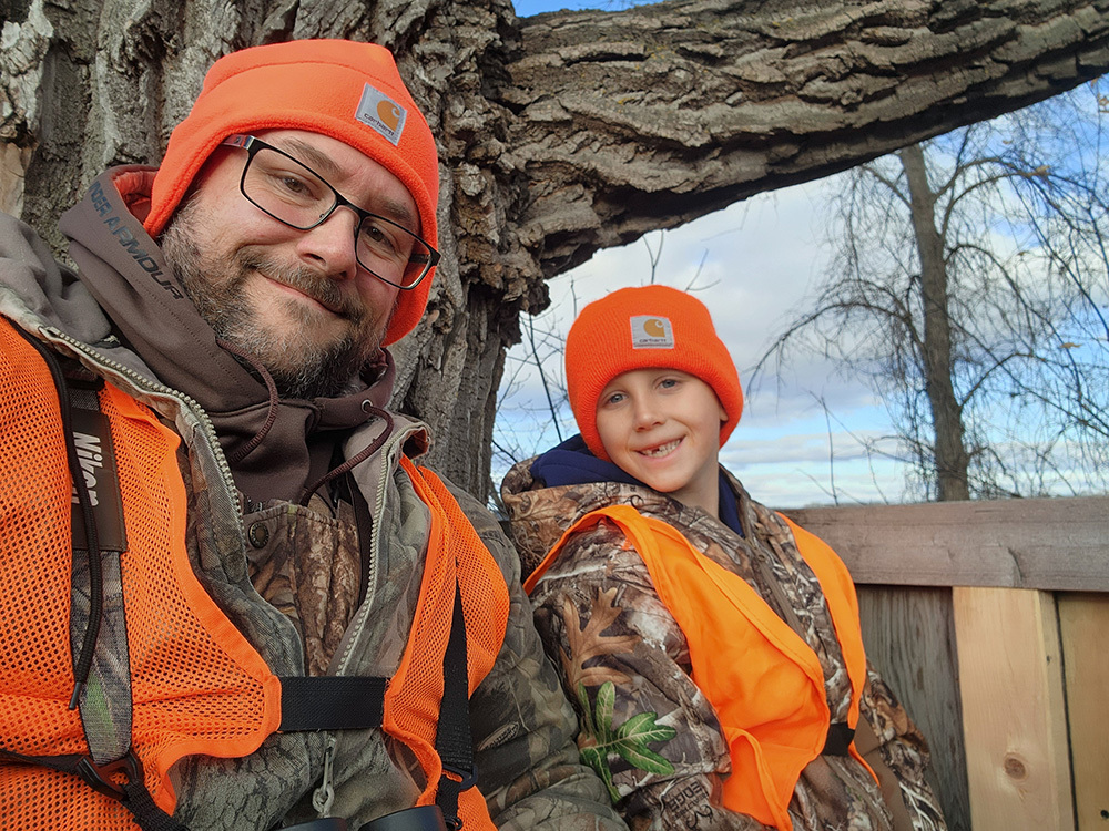 adult and kid out hunting sitting in a tree stand with blaze orange on