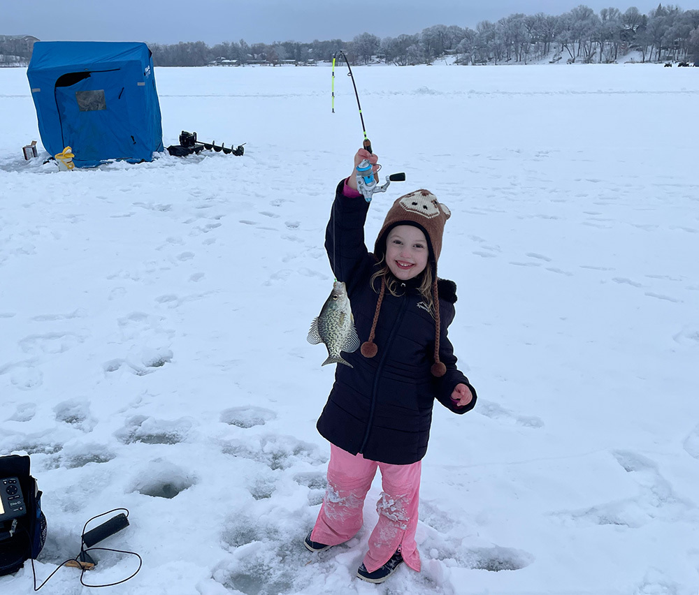 young girl holding up a crappie she caught while ice fishing, hole and portable shelter on ice in foreground and background, respectively