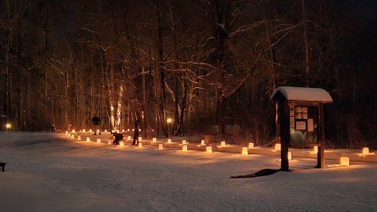 A snowy trail is lined by candles at dusk. 