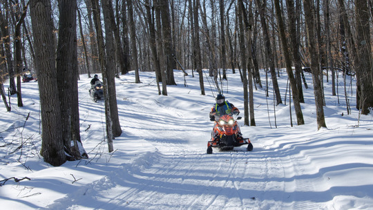 A snowmobile with rider zoom around a corner on a snowy trail through a forest. 
