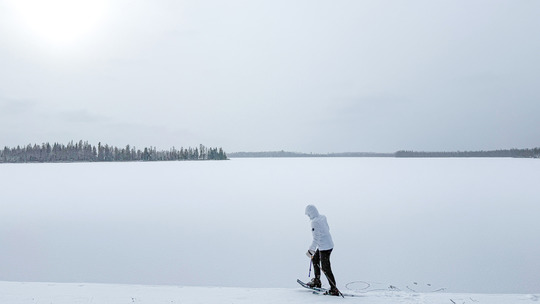 A person in a white jacket snowshoes through a vibrantly white, snowy scene next to a frozen lake.
